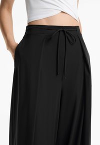 Lockere schwarze Hose mit elastischem Bund, Zugbandsdetail und seitlichen Taschen. Glatte Textur und weit geschnittenes Design.