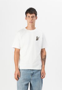 White cotton t-shirt with short sleeves and a round neckline, featuring a butterfly embroidery on the left chest. Casual fit, worn with blue jeans.
