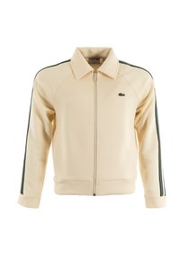 Chaqueta crema con cremallera, con rayas verdes en las mangas, puños y cuello acanalados. Presenta el logo de Lacoste en el pecho. Fabricada con una tela suave.