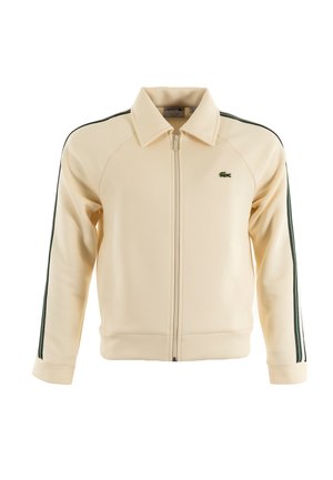 Kremowa kurtka zapinana na zamek z zielonymi paskami na rękawach, żebrowanymi mankietami i kołnierzem. Posiada logo Lacoste na piersi. Wykonana z miękkiego materiału.