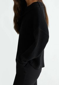 Musta neulepaita, jossa on leveä kaula-aukko, ribbitetty pinta ja tiputetut olkapäät, yhdistettynä slim fit mustiin housuihin.