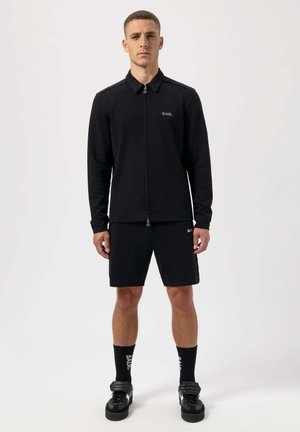 Black zip-up jacket with stand collar and logo, paired with black shorts. Accessories include black socks and sneakers. Simple, athletic design.