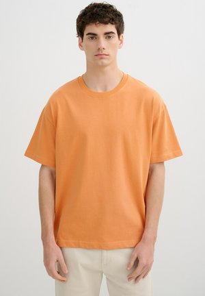 Joven con cabello rizado que lleva una camiseta naranja lisa de manga corta y pantalones beige claro, de pie frente a un fondo gris liso.