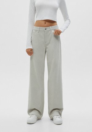 Kobieta ubrana w jasnoszare szerokie spodnie sztruksowe, biały krótki top i białe trampki, stojąca na tle jednolitego jasnego tła.