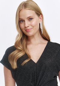 Czarna sukienka z błyszczącymi srebrnymi włóknami; krótkie rękawy, dekolt w kształcie litery V i skręcony detale w okolicy talii. Modelka nosi długie kolczyki w kształcie żyrandola.