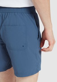 Blaue Shorts aus leichtem Material mit einem elastischen Bund und einer rückseitigen Taschenklappe mit einem Knopf. Glatte, strukturierte Oberfläche.