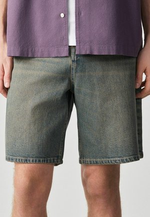Mann trägt ausgewaschene mittelblaue Jeansshorts und ein lila Hemd über einem weißen Oberteil, steht mit entspannt an den Seiten hängenden Händen.