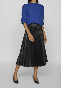 Femme portant un pull bleu roi, une jupe midi plissée noire et des talons slingback à bouts pointus noirs, debout sur un fond gris uni.