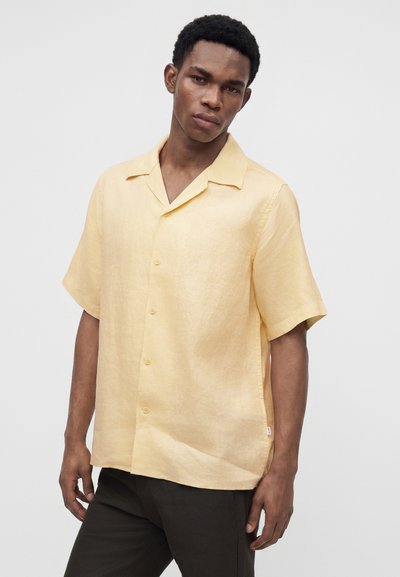 Jeune homme portant une chemise boutonnée jaune clair à manches courtes et un pantalon noir, debout devant un fond blanc uni.