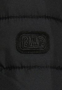 Black, quilted fabric with a textured surface featuring a prominent stitched seam and a raised black "GAP" logo patch.