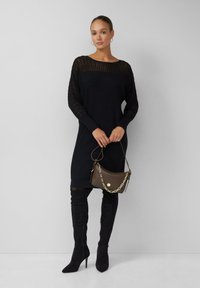 Femme portant une robe noire en tricot côtelé, des bottes noires à talons montant jusqu'à la cuisse, tenant un sac à main brun avec une bandoulière en chaîne dorée devant un fond uni.