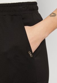 Mano descansando en el bolsillo de unos pantalones negros con una etiqueta de metal cerca del borde del bolsillo, tatuaje de un corazón visible en la muñeca.