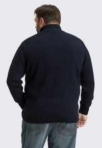 Marineblauwe gebreide trui met een geribde hoge kraag, textuurpatroon op de schouders en geribde manchetten en zoom, gecombineerd met spijkerbroek.