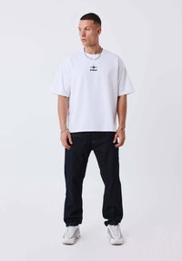 Camiseta blanca de gran tamaño con logo negro, combinada con jeans de mezclilla azul oscuro y zapatillas gris claro. Diseño sencillo, ajuste relajado.