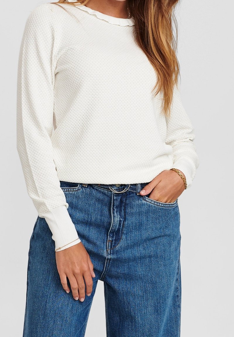 Vit texturerad långärmad tröja med volangkrage, tillsammans med högmidjade blå jeans. Händerna på höfterna med ett armband synligt.