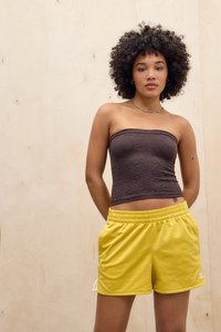 Une jeune femme aux cheveux bouclés porte un haut sans manches à motifs brun et un short jaune Adidas, se tenant devant un fond en bois clair uni.