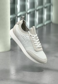 Λευκό και ανοιχτό γκρι αθλητικό παπούτσι με κορδόνια και λογότυπο "ck", εμφανίζεται να αιωρείται πάνω από ένα γυαλισμένο τσιμεντένιο δάπεδο κοντά σε τοίχο από γυάλινα τούβλα.