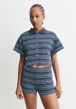 Jonge vrouw met gevlochten haar, gekleed in een marineblauwe en lichtblauwe gestreepte geknoopte crop top en bijpassende high-waisted shorts, staande tegen een witte achtergrond.