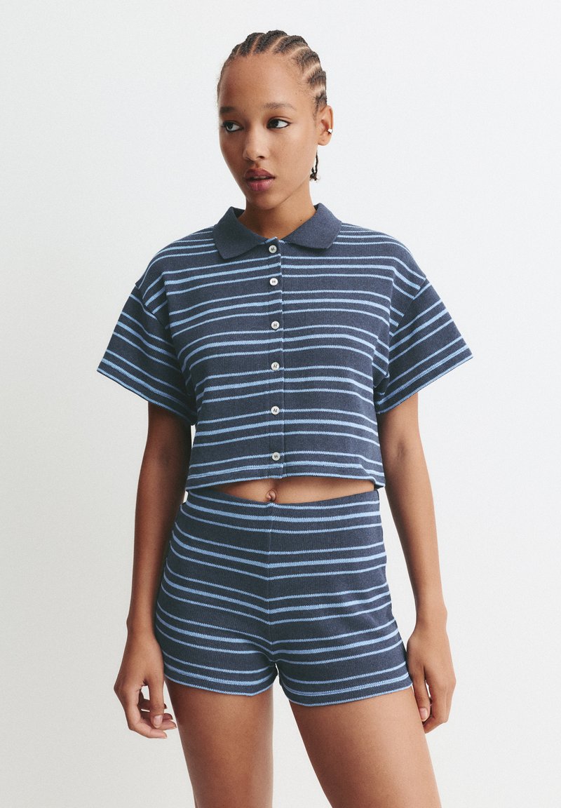 Jonge vrouw met gevlochten haar, gekleed in een marineblauwe en lichtblauwe gestreepte geknoopte crop top en bijpassende high-waisted shorts, staande tegen een witte achtergrond.