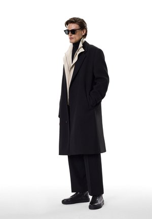 Abrigo de lana negro con interior beige, llevado sobre un suéter de cuello alto negro. Unos pantalones negros y zapatillas completan el conjunto. Los accesorios incluyen gafas de sol negras.