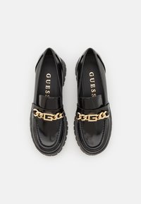 Black patent leather loafers with a golden chain accent and logo, featuring a rugged textured sole for added grip.