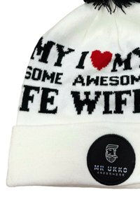 Vit stickad mössa med svart och röd text som säger "MY AWESOME WIFE" och en rund svart etikett med en logotyp och varumärkesnamn. Svart pompomm på toppen.