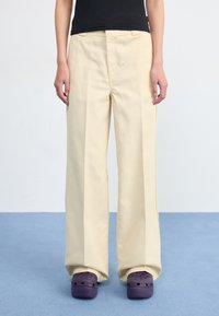 A person stands wearing beige wide-leg pants and purple clogs against a light blue floor and plain white wall.