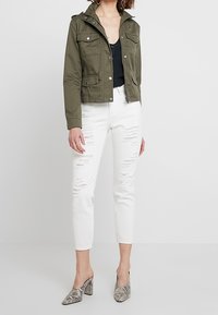 Olivově zelená krátká bunda s kapsami na klopě, bílé distressed džíny a šedé muly s hadím vzorem. Hladká látka, strukturovaný střih, ležérní vzhled.