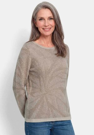 Lachende vrouw van middelbare leeftijd met lang grijs haar, die een beige textuursweater en blauwe spijkerbroek draagt, staand tegen een effen lichte achtergrond.