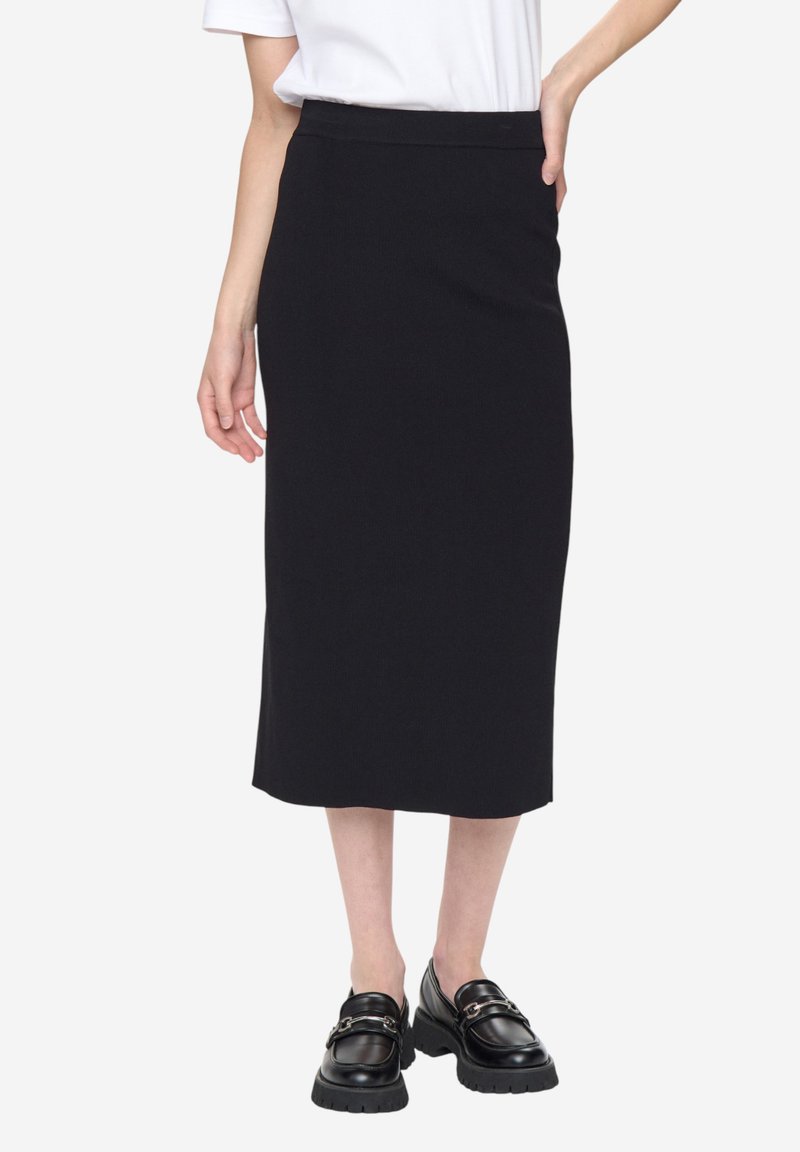 Jupe crayon noire longueur genou en tissu à côtes, avec une ceinture cachée. Associée à des mocassins noirs épais pour un look complet.