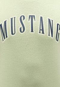 Világoszöld pamut póló, amelyen a "MUSTANG" szó található sötétkék, vastag betűtípussal, fehér kerettel. Az anyag textúrája sima.