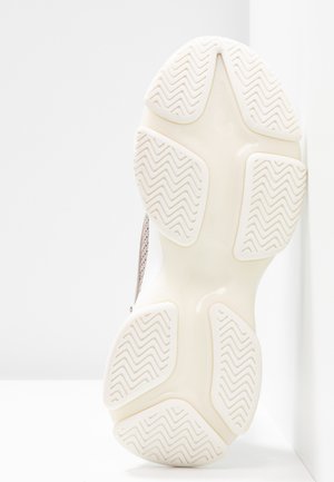 Sole of a light-colored sneaker with textured zigzag tread pattern, positioned upright against a white wall and floor.