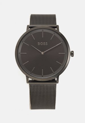 Okrogla črna ura z gladkim številčnico, minimalističnimi oznakami za ure in mrežastim trakcem iz nerjavnega jekla. Logotip "BOSS" je centriran na obrazu ure.