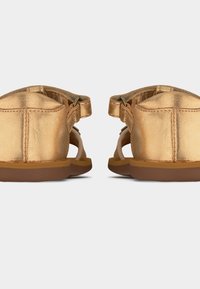 Ζεύγος μεταλλικών χρυσών σανδαλιών από πίσω, με λουριά στον αστράγαλο και καφέ σόλες σε λευκό φόντο.