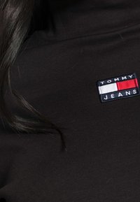 Mustat kangasvaatteet, joissa on suorakulmainen punavalkoinen ja sininen Tommy Jeans -logopatchi olkapään lähellä ja tumma hiushelmi.