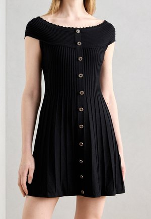 Zwarte geribbelde off-shoulder jurk met verticale plooien en een centrale rij decoratieve ronde knopen op het bovenlichaam van een model.