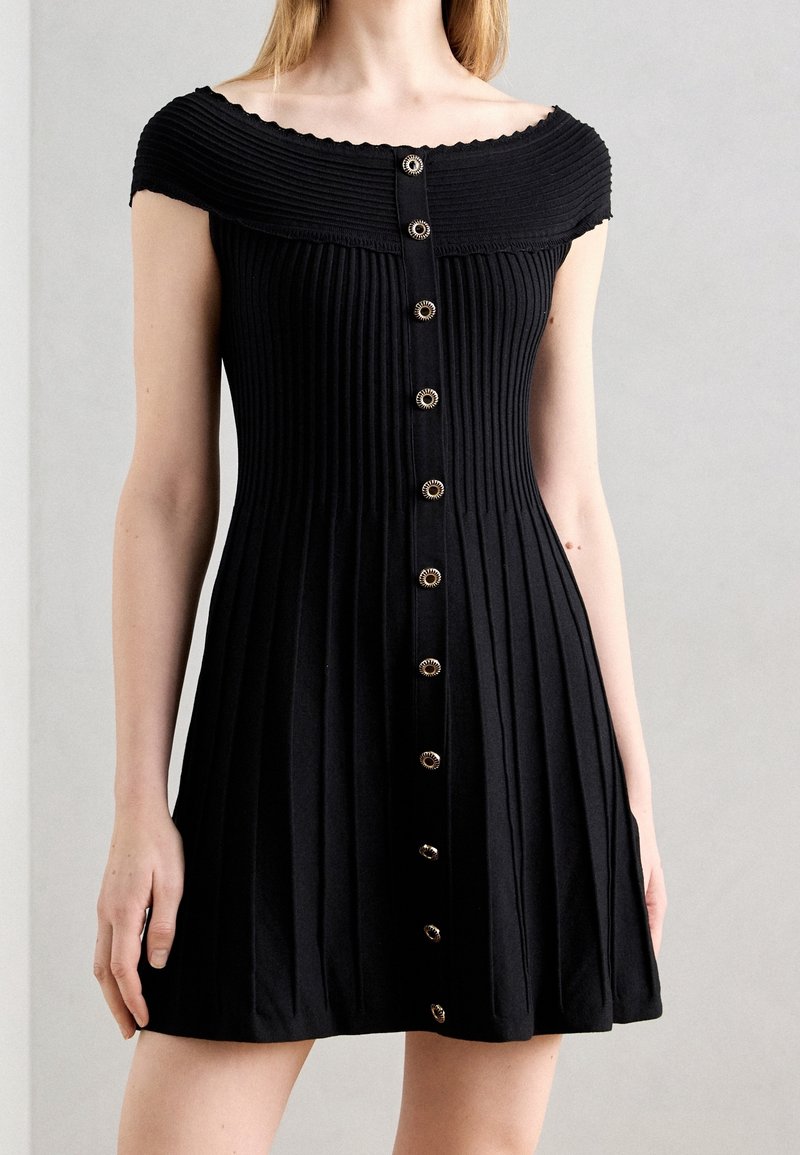 Robe noire côtelée à épaules dénudées avec des plis verticaux et une rangée centrale de boutons ronds décoratifs sur la partie supérieure du corps d'un mannequin.