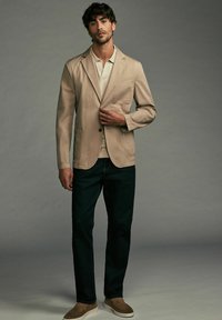 Blazer en coton beige avec un unique bouton, deux poches avant, porté sur un polo clair, associé à un jean foncé et des chaussures slip-on marron.