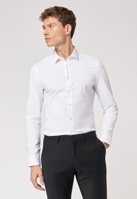 Camicia bianca a maniche lunghe con colletto classico, caratterizzata da una superficie liscia e una leggera lucentezza, abbinata a pantaloni neri su misura.