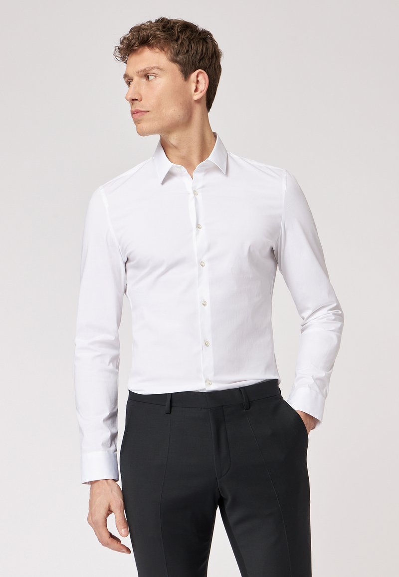 Camicia bianca a maniche lunghe con colletto classico, caratterizzata da una superficie liscia e una leggera lucentezza, abbinata a pantaloni neri su misura.