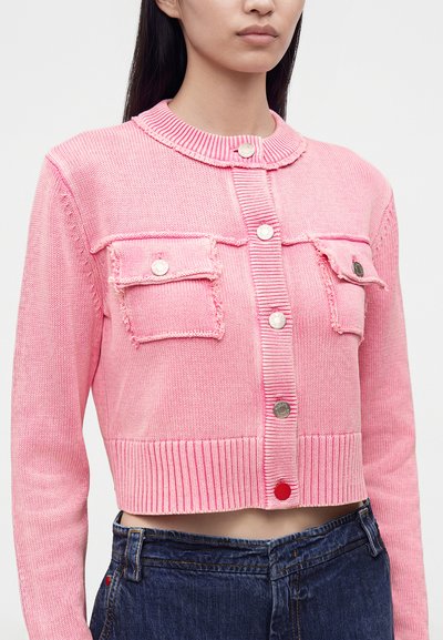 Femme portant un cardigan court en maille rose avec poches avant et boutons argentés, associé à un jean bleu foncé taille haute.