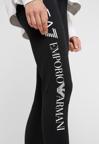 Svarta leggings i stretchmaterial med vit text och logotyp "EMPORIO ARMANI" längs vänster ben. Slät textur, åtsittande design.