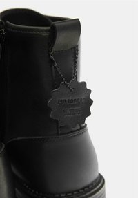 Botte cheville en cuir noir à surface texturée, équipée d'une fermeture éclair latérale et d'une étiquette de marque attachée par une chaîne à perles.