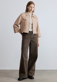 Trumpa smėlio spalvos odinė striukė su struktūruotu apykakle, banguotais rankogaliais ir dviem priekinėmis kišenėmis, derinama su plačiomis rudomis kelnėmis ir juodais batais.