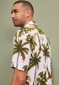 Camicia a maniche corte con bottoni di colore bianco, decorata con un motivo di palme verdi e fiori rossi; tessuto leggero, vestibilità comoda e colletto aperto.