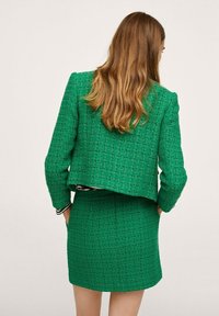 Costume en tweed vert comprenant une veste courte et une jupe ajustée, toutes deux avec un motif texturé et des manches longues. La vue de dos montre un design superposé.
