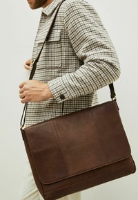 Bolso mensajero de cuero marrón con un acabado texturizado, que presenta una solapa plegable y una correa ajustable. Las costuras salientes añaden detalle.