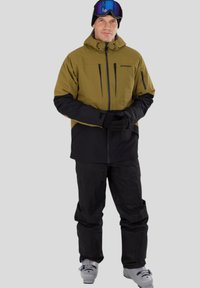 Sjezdová bunda v olivově zelené a černé barvě, s kapucí, zipem a několika kapsami. V kombinaci s černými kalhotami a lyžařskými botami.