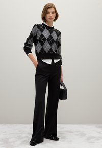 Grijze en zwarte argyle-patroon trui over een witte blouse, gecombineerd met zwarte flared broeken en versierde zwarte instappers, terwijl er een kleine tas wordt vastgehouden.