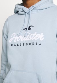 Felpa con cappuccio azzurro chiaro in tessuto morbido, con tasca a marsupio, coulisse e logo ricamato "Hollister California" in bianco e nero.